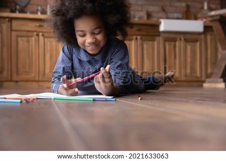Smiling small African American kid lying on floor in kitchen have fun painting drawing in album. Happy cute little biracial ethnic girl child enjoy artistic activity on weekend. Hobby, art concept.