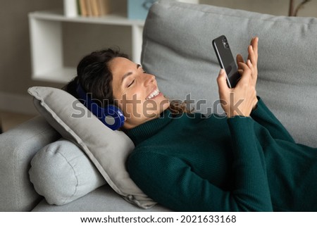 Close up smiling attractive woman in headphones using smartphone, lying on couch, positive young female looking at phone screen, watching video or making call, chatting online, listening music