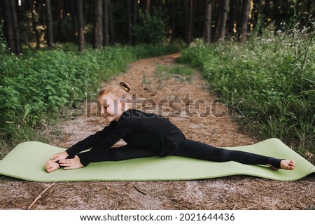 Sports photo of a small, beautiful girl, a child of a professional gymnast in a black silk suit, who diligently trains, warms up before competition in a twine pose in the forest on a green rug.