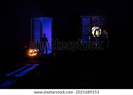 Halloween horror concept with glowing pumpkin. A realistic dollhouse living room with furniture, door and window at night. Scary zombies outside. Selective focus.