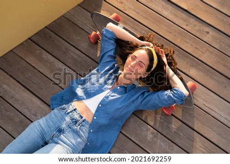 Cool trendy young girl lying on longboard with eyes closed wearing stylish clothes and hipster eyeglasses at sunset. Overhead view of hippie female skateboarder on wooden deck relaxing on skateboard