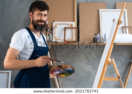 close-up portrait of a bearded male artist in a dirty apron stands in an art studio and looks into the camera. holds in his hands a palette with different colors of paints.
