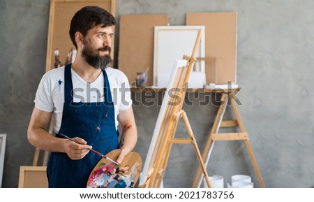 A bearded male artist dressed in a blue dirty apron stands in an art studio holding a palette with paints in his hands looks at the empty space for the text.