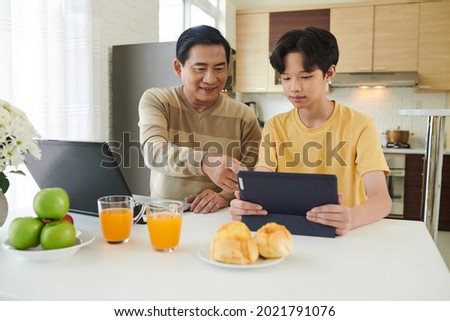 Father and son working at home next to each other, they are sitting at kitchen table with laptop and digital tablet Royalty-Free Stock Photo #2021791076