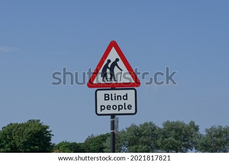 A red and white triangle warning sign and Blind People notice against a blue sky with an image of elderly fragile people advising drivers to be aware of people crossing the road
