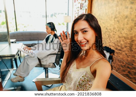 Young female attractive millenial Asian mixed European race youtuber waving at audience online while holding a camera at barbershop salon.