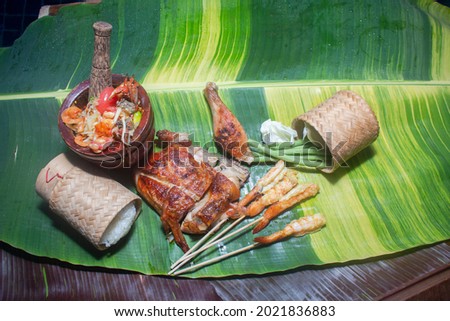 Sticky rice, papaya salad, grilled chicken and grilled shrimp served on a spotted banana leaf poolside.
red yellow green speckled banana leaves in the background. food and restaurant concept.
