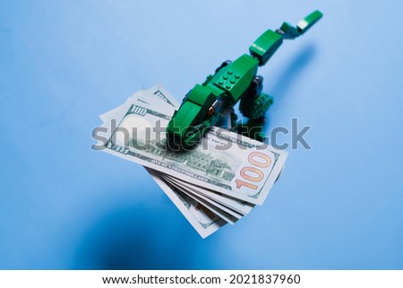 A pack of 100 dollar bills in mouth of tyrannosaur green kid toy isolated on blue background. Copy space. Concept of a unique advertising and design.