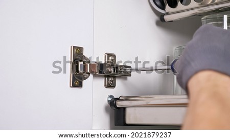 Skilled employee hands in grey textile gloves install door of white cabinet twisting screws in hinges in kitchen extreme close view