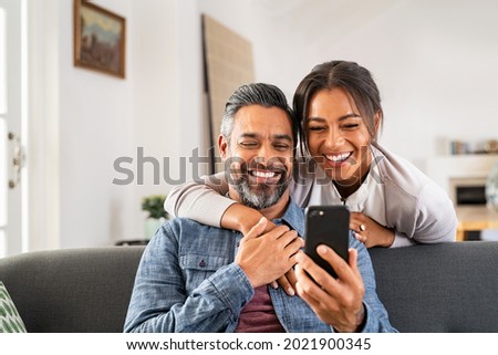 Mid adult man relaxing on sofa and showing new app to african american wife on cellphone. Middle eastern man and woman sitting on couch at home and using mobile phone to do a video call with family. Royalty-Free Stock Photo #2021900345
