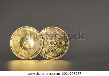 two cardano cryptocurrencies standing side by side overlapping one for each side of the coin glowing with side light and gray background with gold reflection in front Royalty-Free Stock Photo #2021908937
