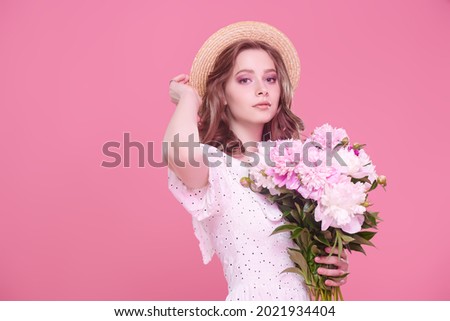 A pretty girl in a light white summer dress and a straw hat stands with a bouquet of peonies on a pink background. Summer beauty and fashion. Happy people.
