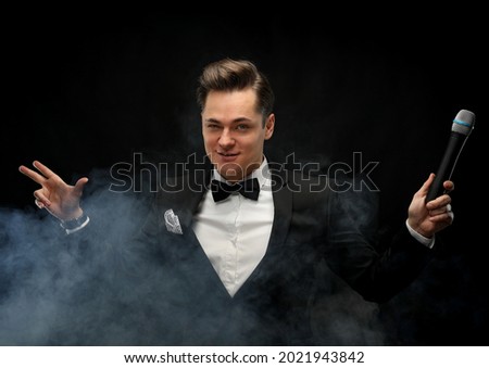 Stylish young man in a tuxedo holding a microphone, posing against a dark background with smoke, actor, singer, show, host of the event.