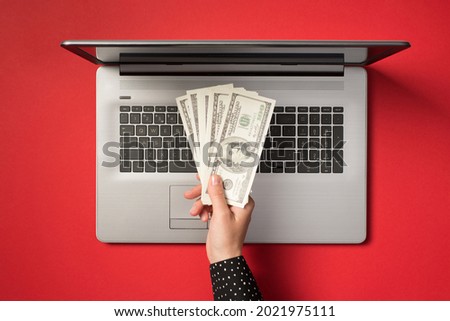 Overhead photo of grey laptop and hand holding a pile of hundred dollars isolated on the red backdrop