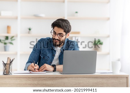 Positive arab male manager working from home during COVID-19 pandemic, using modern laptop and taking notes, sitting at workdesk in living room interior, copy space