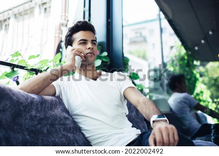 Handsome male generation Z in casual clothes calling to customer service for discussing details using wireless connection at terrace, millennial hipster guy using cellular technology for phoning