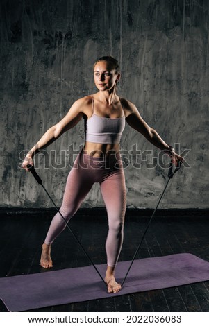 Vertical portrait of muscular young athletic woman with perfect beautiful body wearing sportswear exercising with resistance band. Caucasian fitness female training with stretching expander in studio.