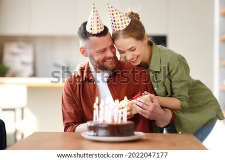 Young positive wife making surprise for husband, excited man holding gift box and being so happy while sitting in kitchen at table with cake. Family couple in party hats celebrating birthday at home