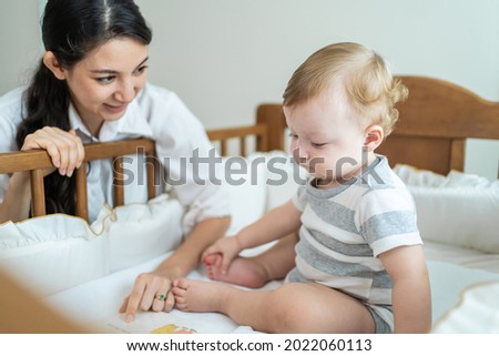 Caucasian mom read storytale to baby infant in child crib in bedroom. Beautiful parent, mother play education game picture book with happy little young boy son kid on bed in house. Parenting activity.