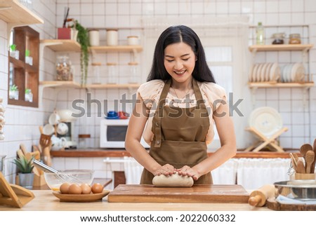 Asian beautiful mature woman stay home, spend time in kitchen cooking. Young attractive carring female wear apron and smile, kneaded yeast dough with hand to baking bakery on table for dinner in house Royalty-Free Stock Photo #2022060332