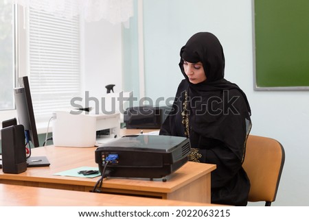 A Muslim teacher is sitting at her desk in a school classroom.