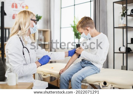 Caucasian boy in medical mask sitting at hospital cabinet while competent doctor making injection in shoulder. Concept of vaccination, medicine and health care.