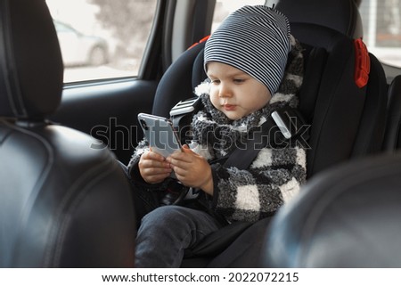 Toddler sits in a child car seat and watches cartoons, holds a mobile phone. The boy is dressed in an autumn sweatshirt and a hat. Little kid 2-3 years old plays a game on gadget 