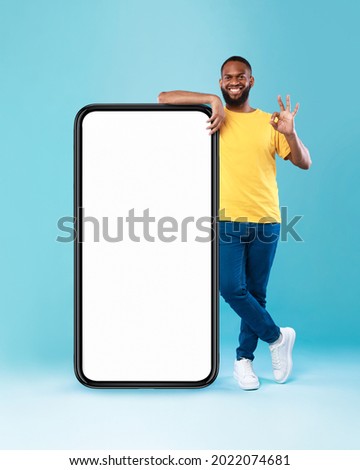 Positive black guy showing okay gesture next to huge smartphone on blue studio background, empty space for mobile app design on screen. Website or advertisement mockup