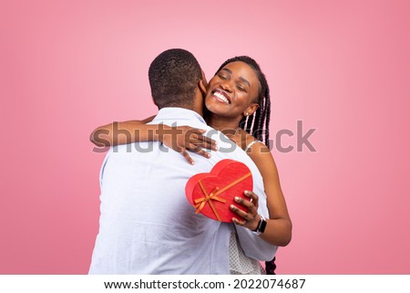 Celebrating Special Occasion. Portrait of romantic African American guy giving his girlfriend wrapped box with present in heart shape and hugging, man greeting lady with holiday, pink studio wall