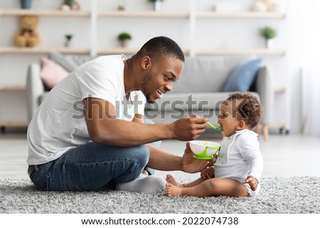 Father's Care. Loving Black Dad Feeding His Cute Baby Son From Spoon At Home, Young African American Daddy Giving Healthy Food To His Little Toddler Child While They Relaxing Together In Living Room Royalty-Free Stock Photo #2022074738
