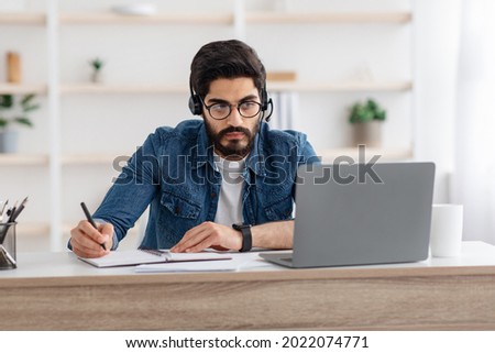 Millennial arab guy in headset watching online webinar on laptop at home, having online training, studying with computer and taking notes to notepad, copy space