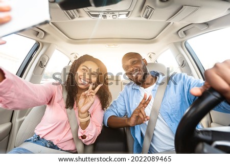 Happy black lovers taking selfie while having car trip, sitting in auto, cheerfully smiling at camera, pretty millennial girlfriend and boyfriend showing v victory peace sign gesture, sun flare