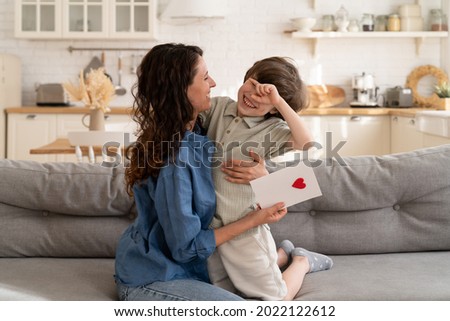 Family celebration: small kid presenting mom greeting card with heart on mother day, birthday or valentines holiday. Cute boy child congratulating mum. Happy parent embrace loving son on festive event