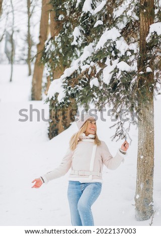 Beautiful girl in a Winter Suede Lambs Wool Jacket enjoying winter moments. Outdoors photo of blonde in a pink hat having fun on a snowy morning on a blurred nature background.