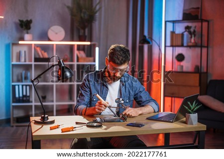 Handsome 35-aged bearded man in protective glasses sitting at table and soldering video card from modern laptop. Young male repairing important detail from computer by himself at home.