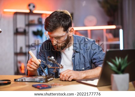 Young man with beard fixing display card from laptop using soldering iron. Handsome guy wearing protective glasses while repairing detail from computer. Royalty-Free Stock Photo #2022177776