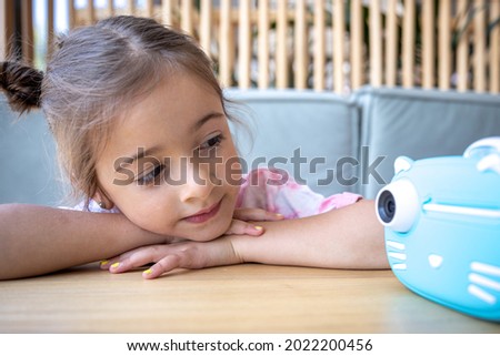 A cute little girl looks at her children's blue toy camera for instant photo printing.