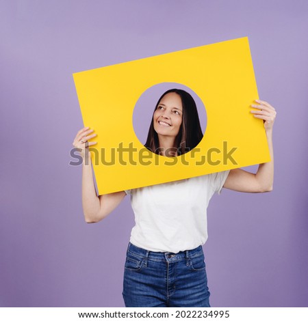 Positive woman looking up through a cutout in a yellow board with a smile of hope as she seeks inspiration over a purple studio background with copyspace in square format