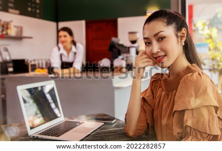 Portrait beautiful Asian businesswoman is a sitting customer at the desk with laptop computers inside the coffee shop Wi-Fi Internet cameras pleasantly welcomed by a female barista at the counter.