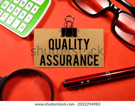 Business concept.Text QUALITY ASSURANCE writing on brown card with magnifying glass,glasses,calculator and pen on red background