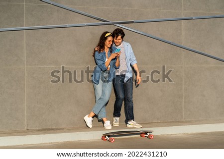 Young trendy couple use smartphone together connecting to wireless 5g internet outdoors at modern urban space. Happy longboarders man and woman sharing video or photo in social media in mobile phone