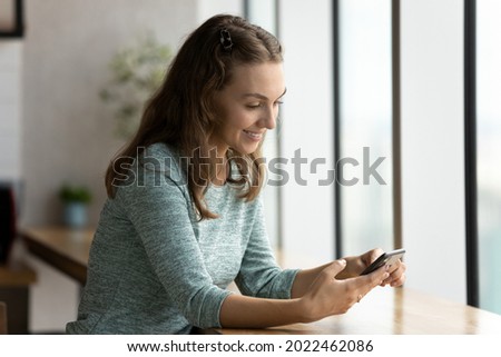 Happy gen Z girl with smartphone receiving good news, reading text on screen. Young woman using app on mobile phone at desk, shopping, paying for purchase, chatting on social media, dating website