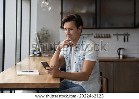 Happy thoughtful millennial guy using app, online service on smartphone at home workplace table, looking at screen, browsing internet, reading text, typing message. Man making call on mobile phone