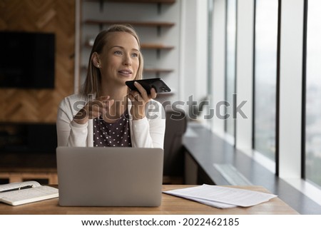 Happy business woman recording audio message on cellphone at workplace, making phone call on speaker, giving audio command to virtual assistant for online search, using voice recognition app