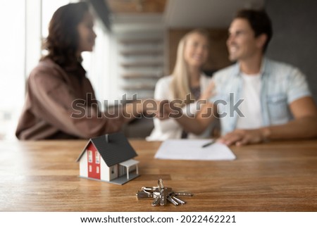 Close up of tiny toy house model and key on office table. Married couple of clients and real estate agent giving handshakes in office. Customers meeting with broker, seller for signing agreement Royalty-Free Stock Photo #2022462221