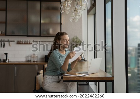 Happy young woman reading paper letter at home workplace with laptop, getting good news, bank or insurance notice. Gen Z student girl receiving approved application from college or university Royalty-Free Stock Photo #2022462260