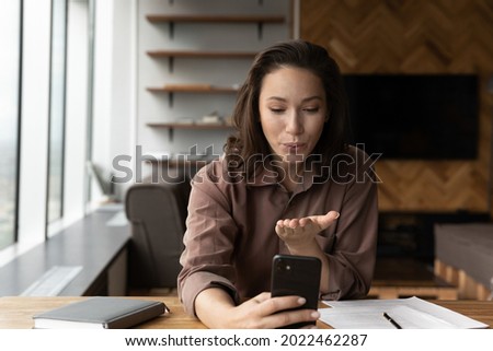 Happy young business woman making personal video call from workplace, holding smartphone, sending air kiss to screen. Female employee using work break for private conference talk, taking selfie
