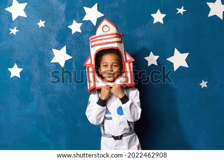 Rocket afro-american little boy in space suit playing astronaut on blue background with white hand-made stars. Happy mixed race child with handcrafted spacecraft. Childhood, creative and imagination. Royalty-Free Stock Photo #2022462908