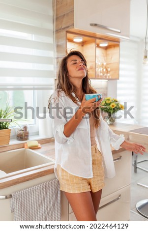 Beautiful young woman standing in the kitchen, drinking cup of hot coffee, relaxing at home in the morning
