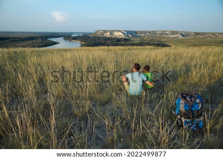 Dad hugs his son, sitting on the grass on the hillside and look into the distance, into the future, with a tourist backpack lying on the grass